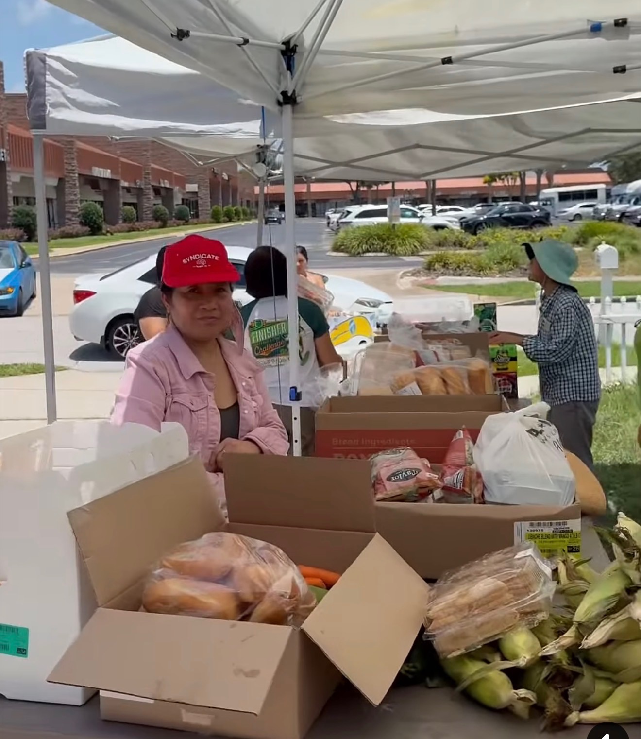 A volunteer in a pink jacket and red cap standing behind a table of fresh produce, bread and corn at a Hands to the Needy distribution in Orlando